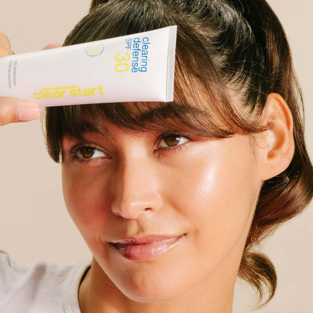 Woman holding a tube of 'Clear Start' sunscreen with SPF 30 against a neutral background